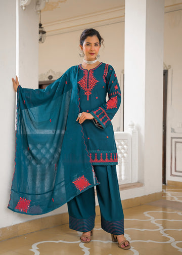 A woman models a teal blue ethnic three-piece suit with red embroidery, featuring a long kurta, wide-leg pants, and a matching dupatta, posed in an elegant indoor setting with white pillars.