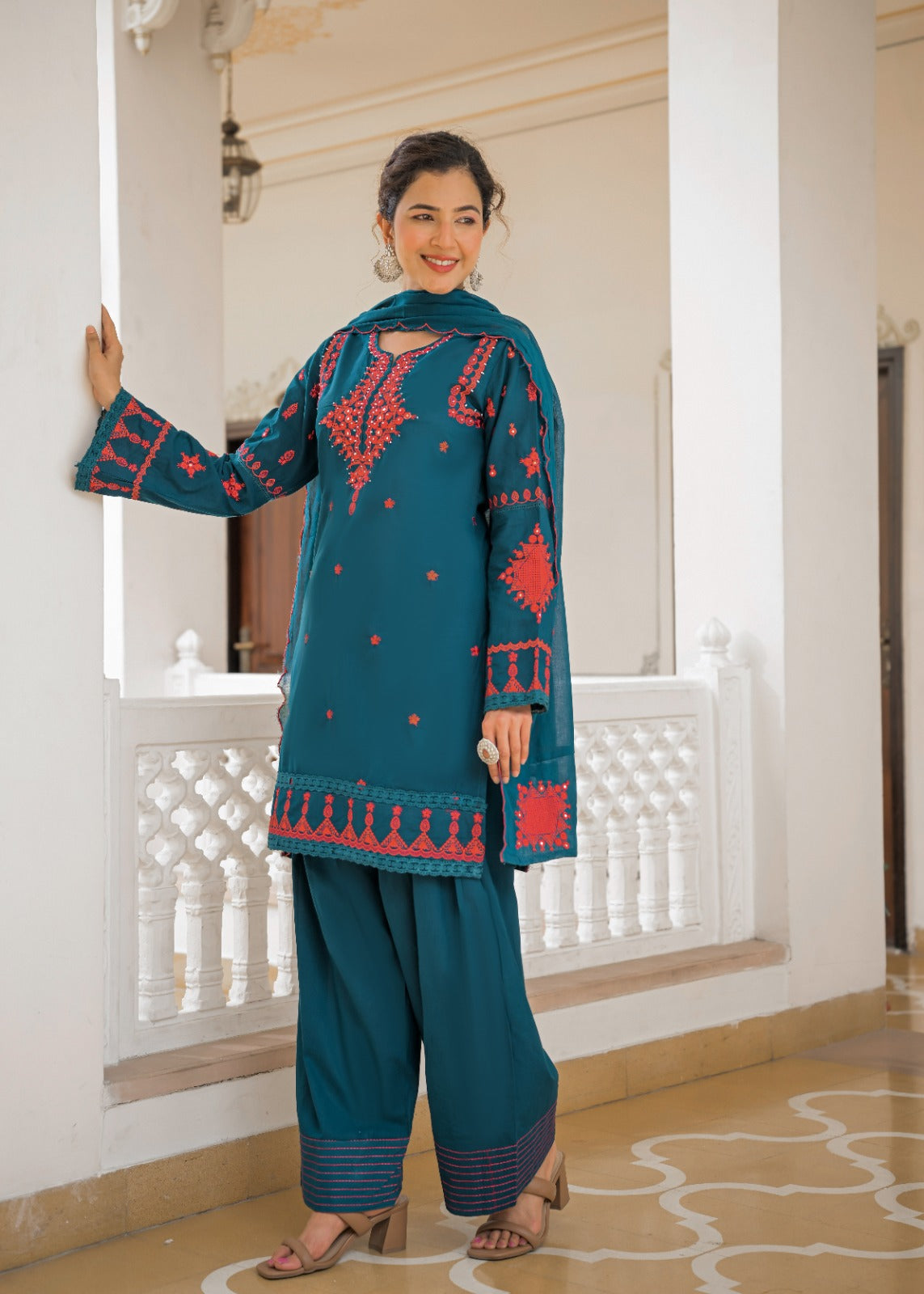 A woman modeling a teal blue ethnic salwar kameez suit with red embroidery, standing in a white-walled indoor corridor with a decorative railing.