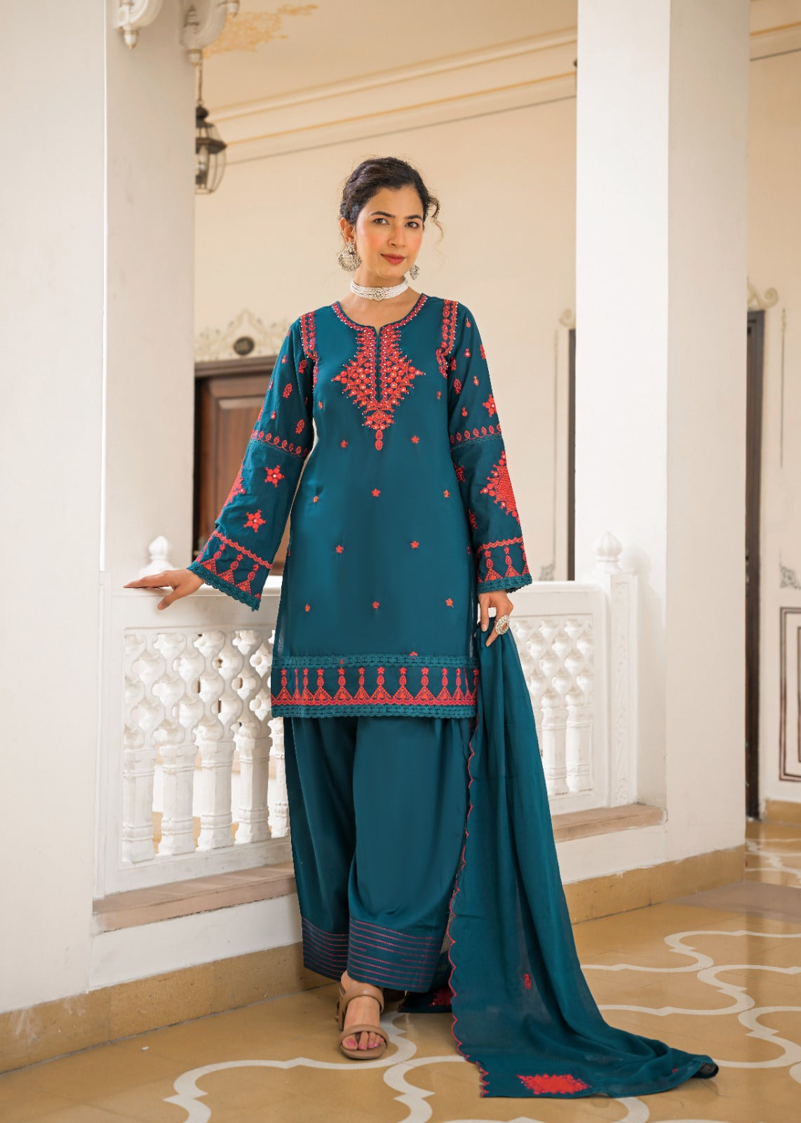 A woman models a traditional South Asian salwar kameez suit in teal blue with red and gold embroidery, paired with matching wide-leg trousers and a long dupatta, set against an elegant indoor architectural background.