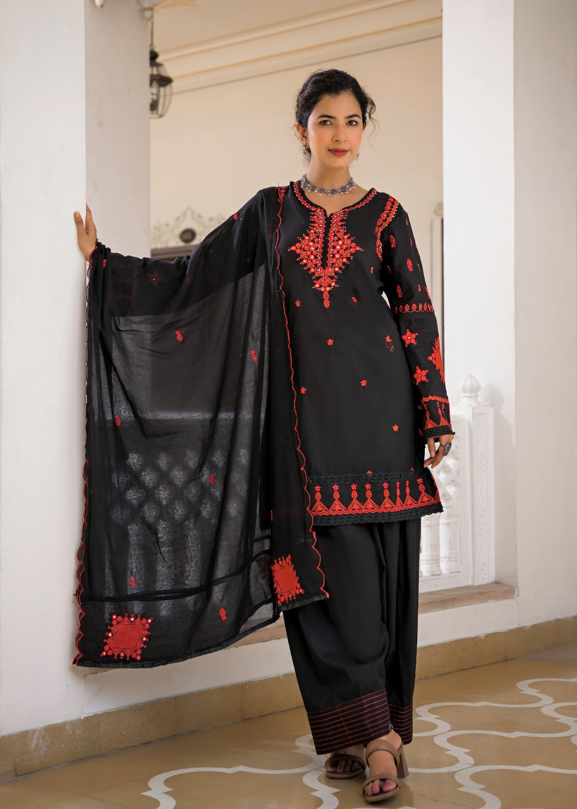 A woman models a black traditional South Asian salwar kameez suit with red embroidery and a matching black dupatta in an indoor setting with white pillars.