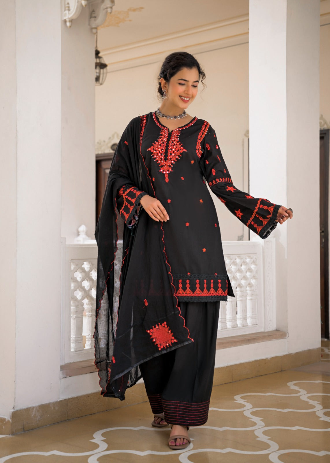 A woman modeling a black traditional salwar kameez suit with red embroidery, featuring a long kurta, matching trousers, and a sheer dupatta, set against an elegant indoor corridor with white pillars.