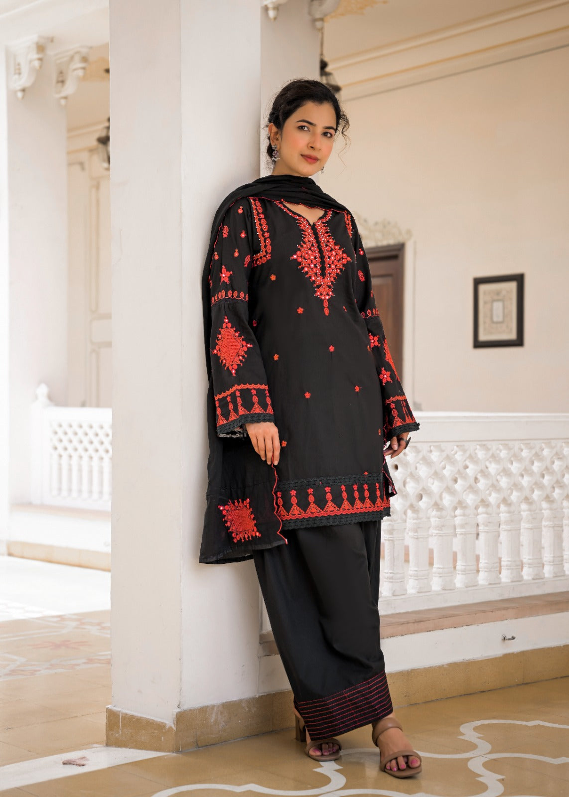 A woman models a black traditional kurta set with red embroidery, consisting of a long-sleeve tunic and matching wide-leg trousers, in an elegant indoor corridor.