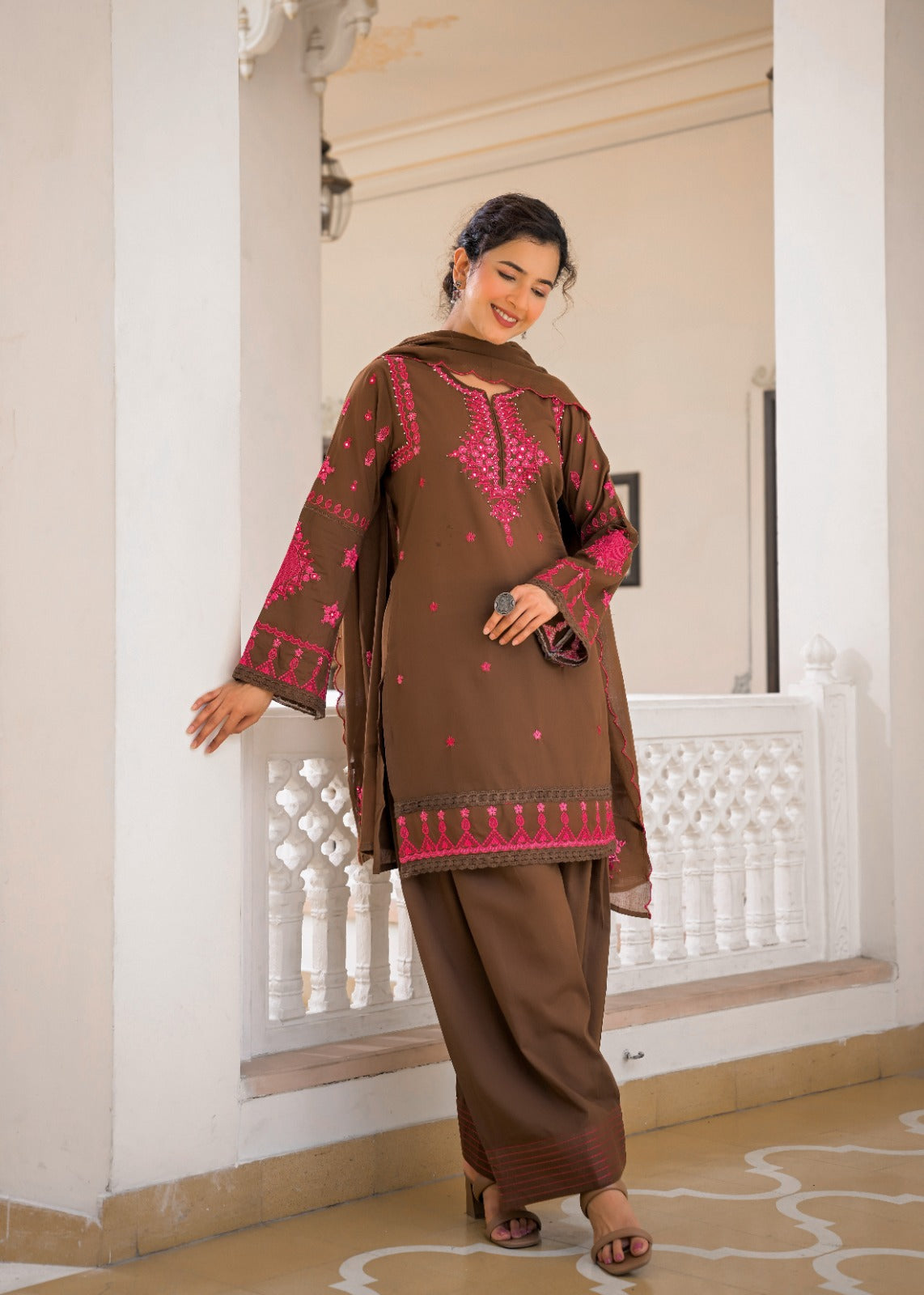 A woman models a brown three-piece salwar kameez suit with vibrant pink embroidery, standing in a white-walled corridor with a white railing.