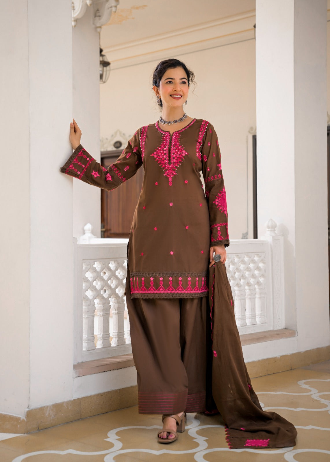 A woman models a brown ethnic salwar kameez suit with vibrant pink embroidery, featuring a long kurta, matching wide-leg pants, and a coordinating dupatta, set against an elegant white architectural background.