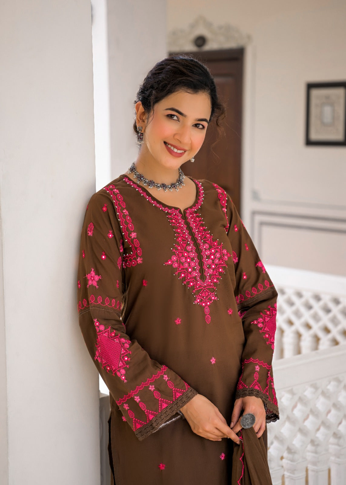 A woman wearing a brown long-sleeve kurta with vibrant pink and silver embroidery, leaning against a white wall in an indoor setting.