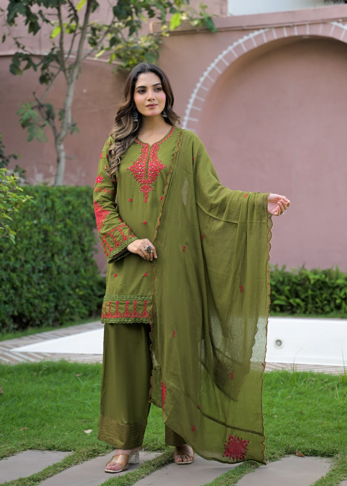 A woman stands outdoors in a garden wearing an olive green traditional three-piece salwar kameez suit with red floral embroidery on the kurta and trousers, paired with a matching sheer dupatta.