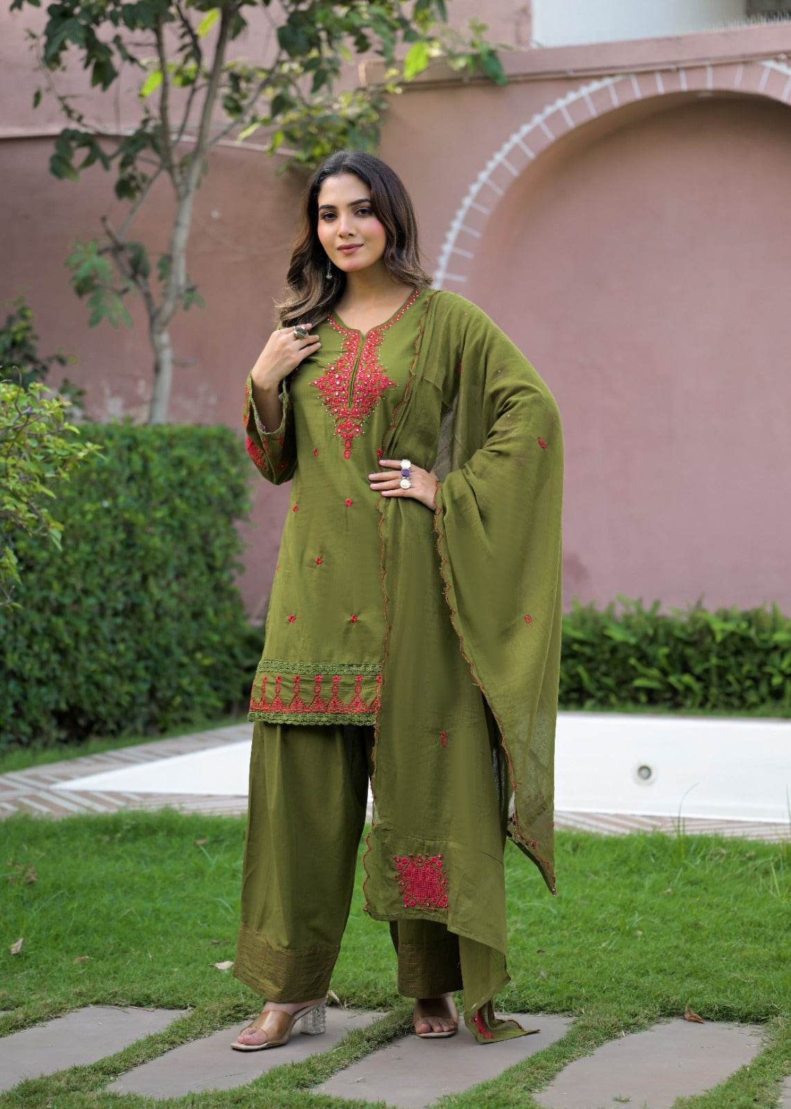 A woman stands outdoors on a stone path wearing an olive green traditional outfit with red embroidered details and a matching draped scarf. The setting includes green grass, shrubs, and a pinkish stucco building in the background.