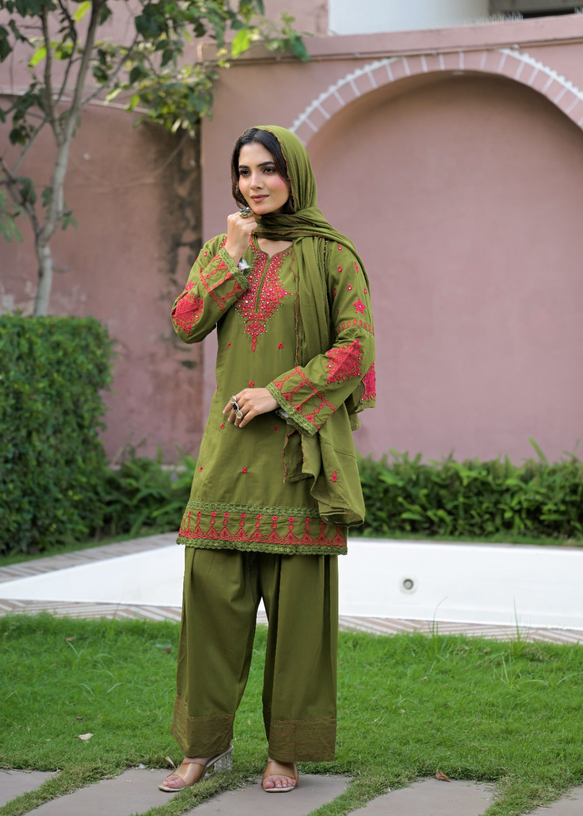 A woman modeling an olive green three-piece salwar kameez suit with red embroidery, matching olive green trousers and dupatta, standing outdoors on a paved path with a garden and pinkish wall in the background.