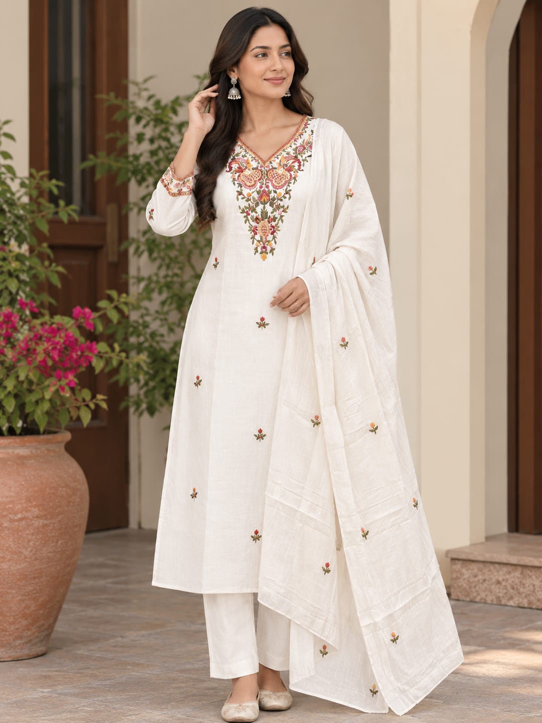 A woman models a white traditional Indian salwar kameez suit with intricate floral embroidery around the V-neckline and scattered embroidered motifs on the kurta and dupatta, set against an elegant outdoor patio backdrop.