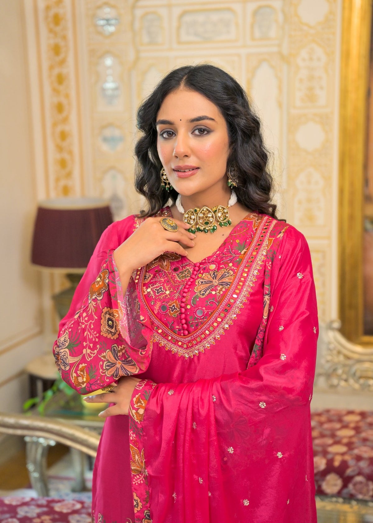 A woman wearing a bright pink traditional embroidered outfit with ornate gold and multicolor detailing, standing in an ornate indoor setting with decorative walls and furniture.