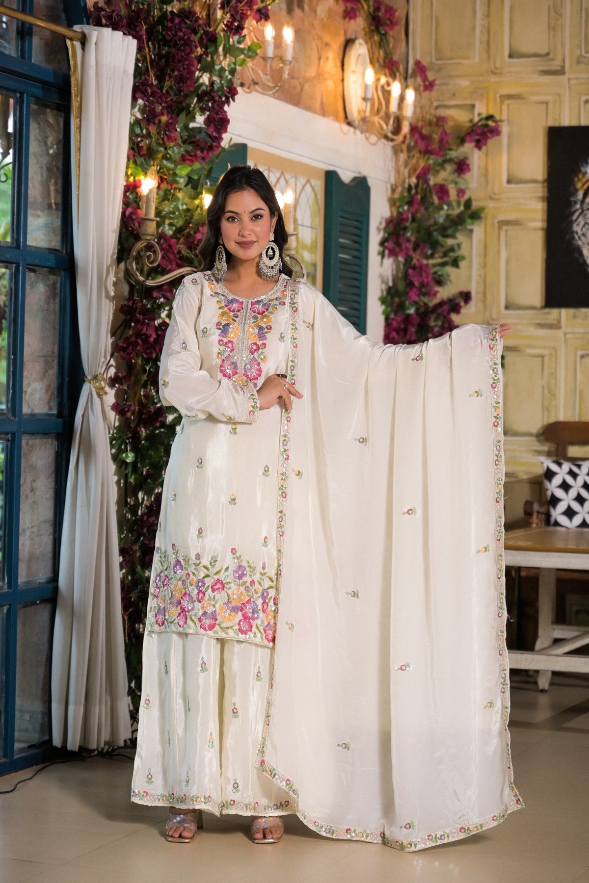 A woman models a traditional white Indian salwar kameez suit with colorful floral embroidery and a matching dupatta, posed in an ornate indoor setting with floral decorations.