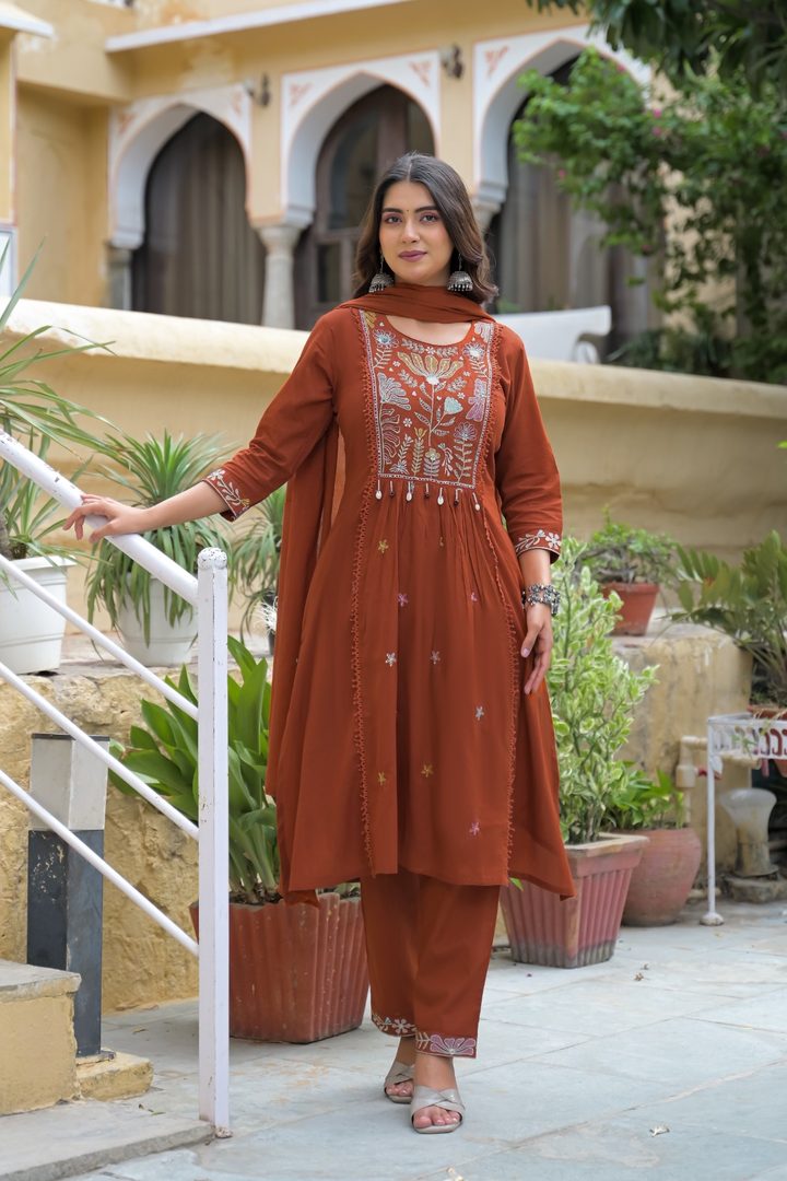 A woman modeling a rust-brown kurta and trouser set with white embroidery, standing in an outdoor courtyard setting with potted plants and a beige building in the background.