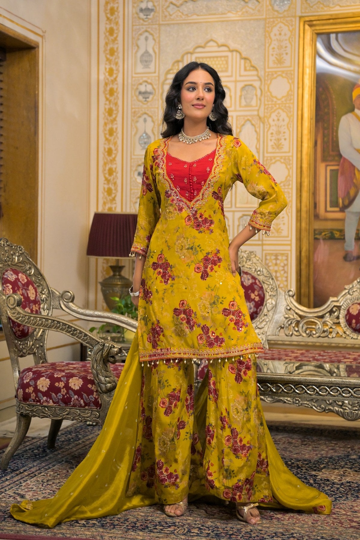 A woman models a traditional Indian salwar kameez suit in a richly decorated room. The outfit consists of a mustard yellow embroidered kameez jacket with a red inner bodice and matching yellow palazzo pants, accompanied by a sheer yellow dupatta.