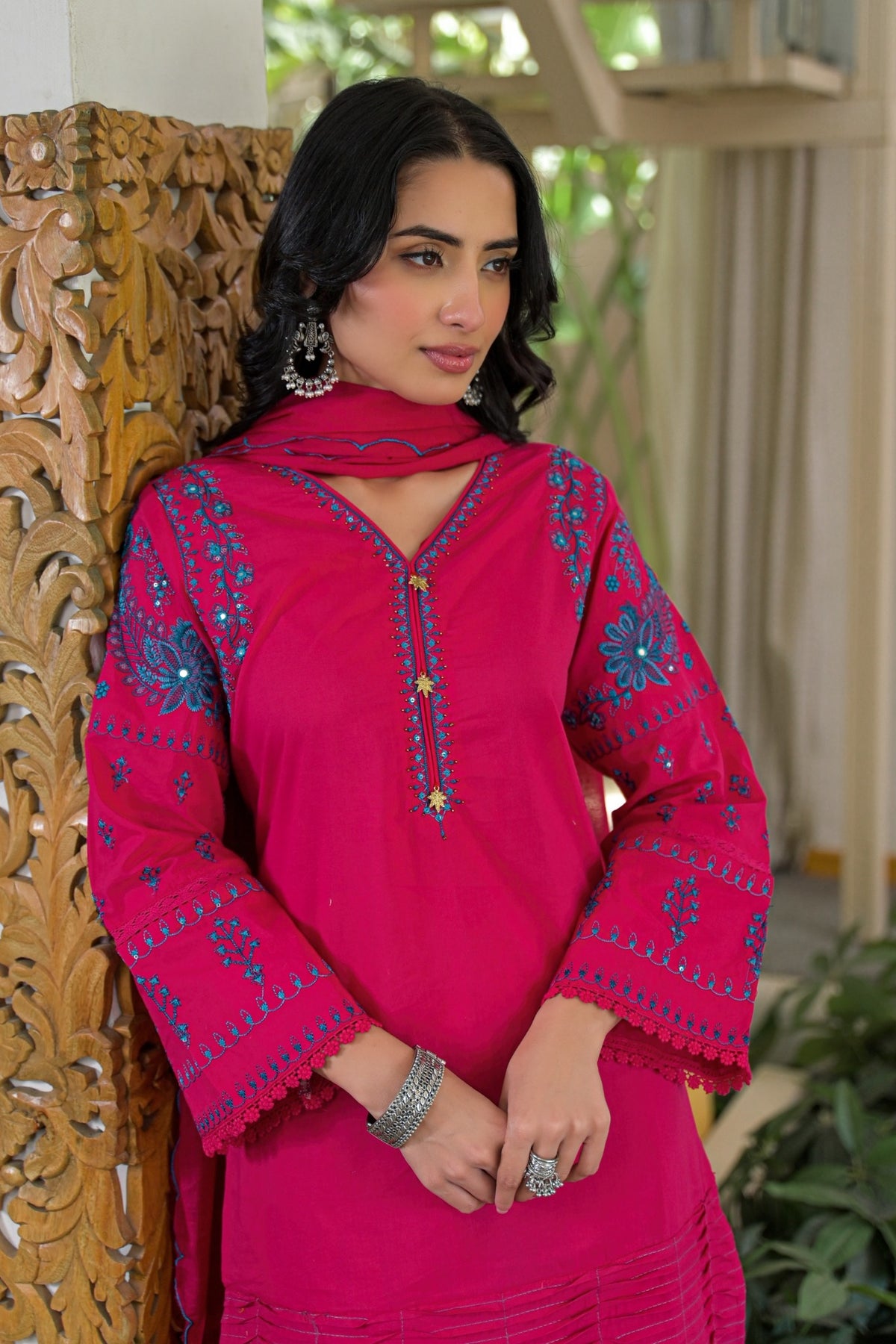 A woman wearing a bright pink embroidered kurta set with a matching dupatta, standing in front of a carved wooden panel.