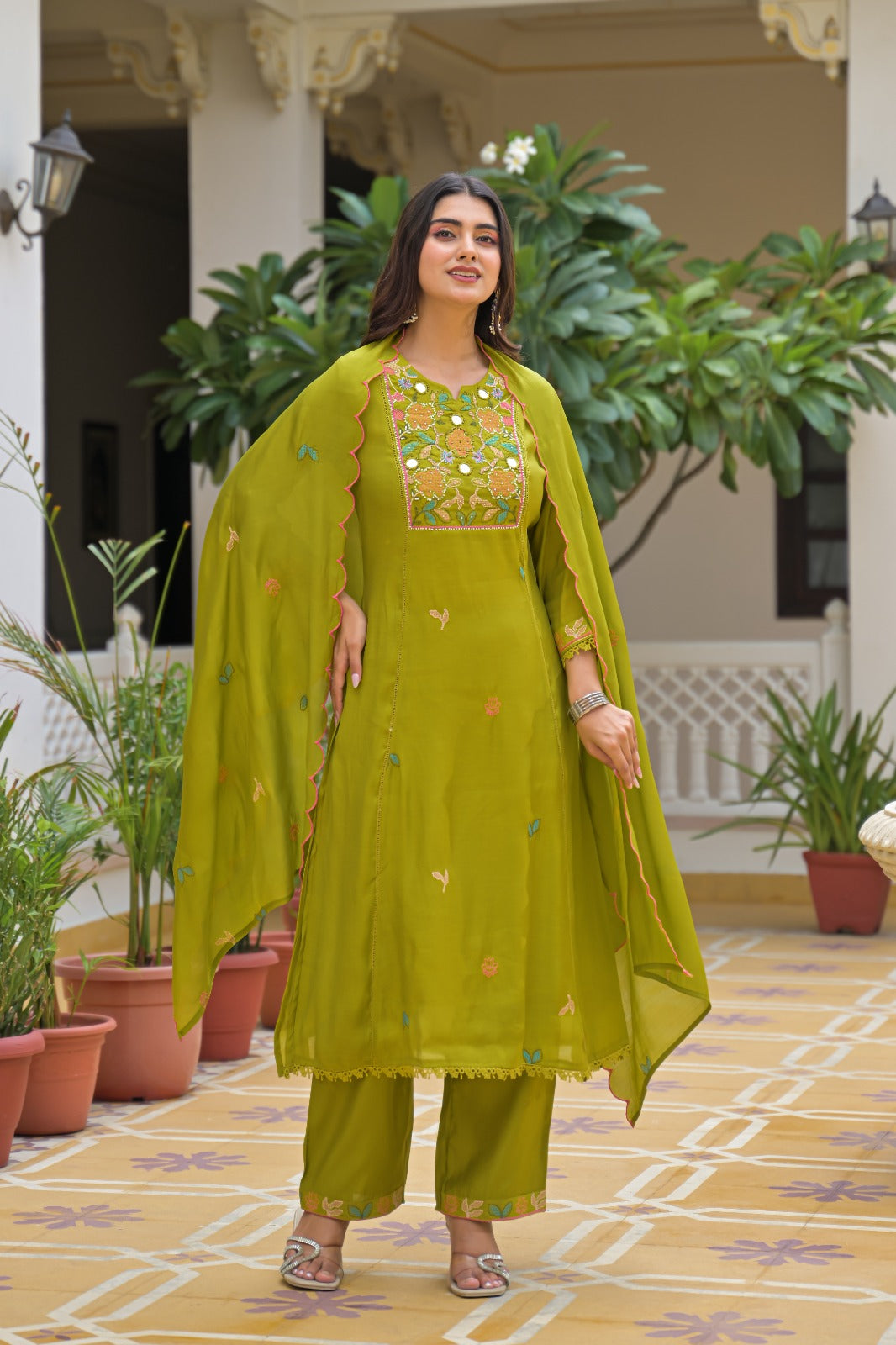 A woman stands in a courtyard wearing a bright olive green traditional outfit with a matching dupatta. The outfit includes an embroidered kurta with a decorative neckline, matching pants, and small embroidered motifs.