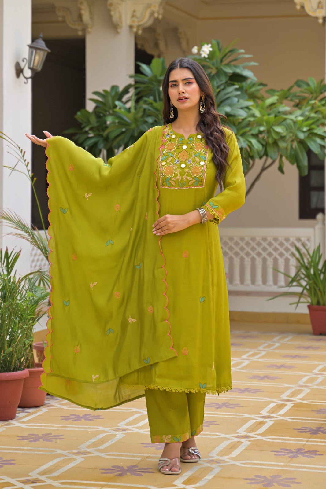 A woman models a traditional Indian salwar kameez suit in olive green, featuring a printed kameez top with colorful floral embroidery, matching green pants, and a sheer green dupatta with embroidered details, set against an outdoor courtyard background.