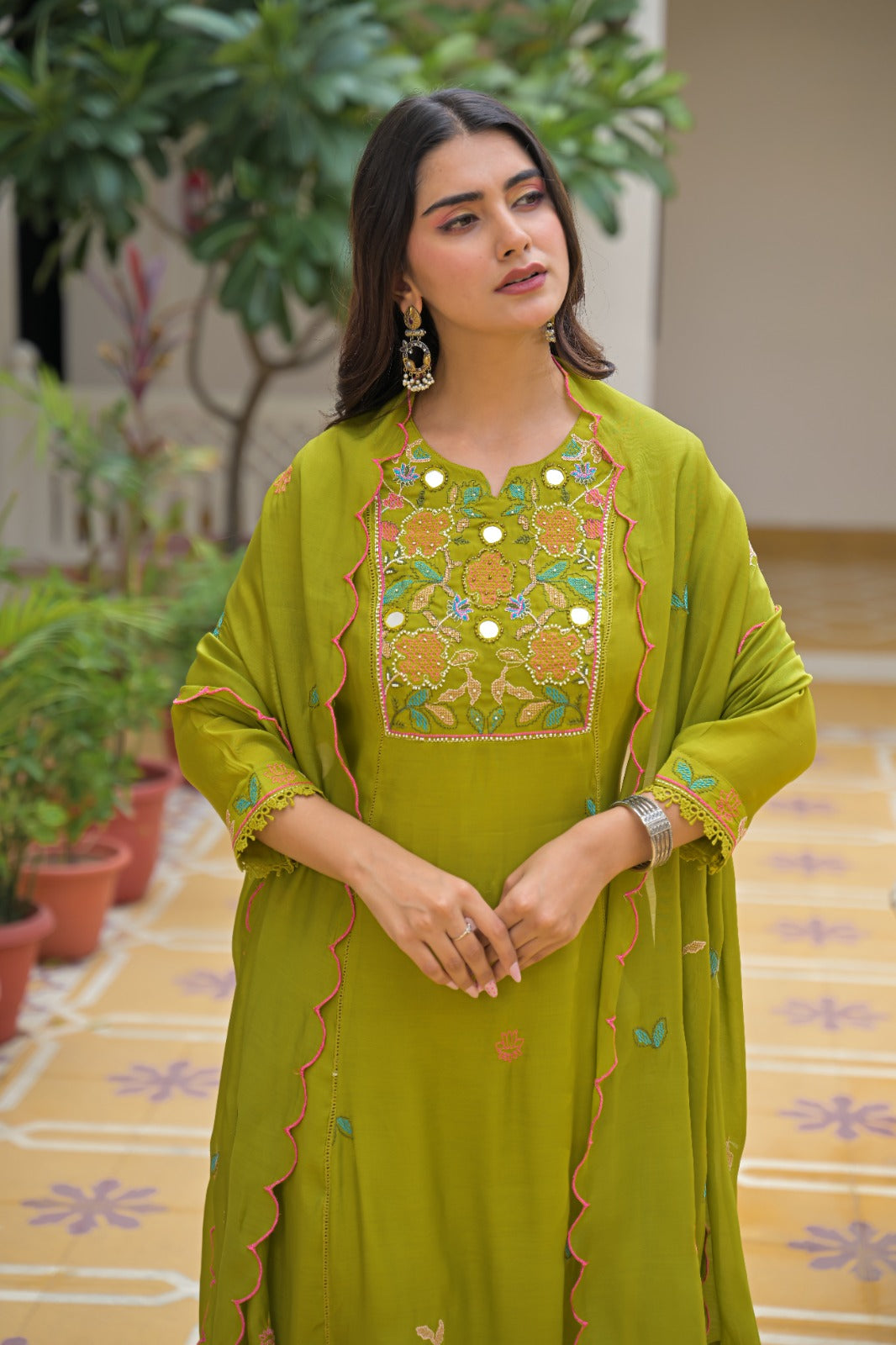 A woman stands in a courtyard wearing a bright olive green traditional outfit with a matching dupatta. The outfit includes an embroidered kurta with a decorative neckline, matching pants, and small embroidered motifs.