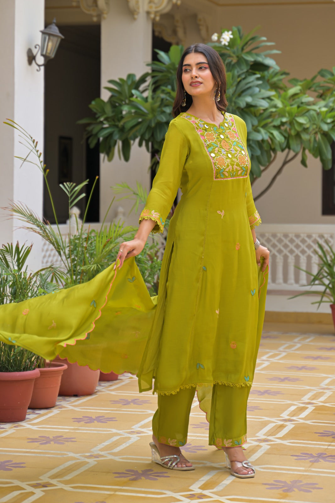 A woman models a bright yellow-green salwar kameez suit with matching dupatta in an outdoor courtyard setting. The kurta features a colorful floral embroidered neckline and small embroidered motifs on the front, paired with matching pants and a sheer dupatta.