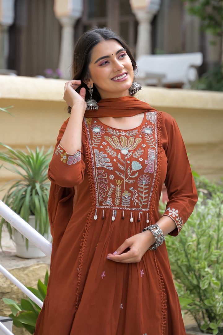 A woman wearing a rust-orange embroidered kurta with a matching dupatta, standing outdoors in front of a beige wall and green plants.