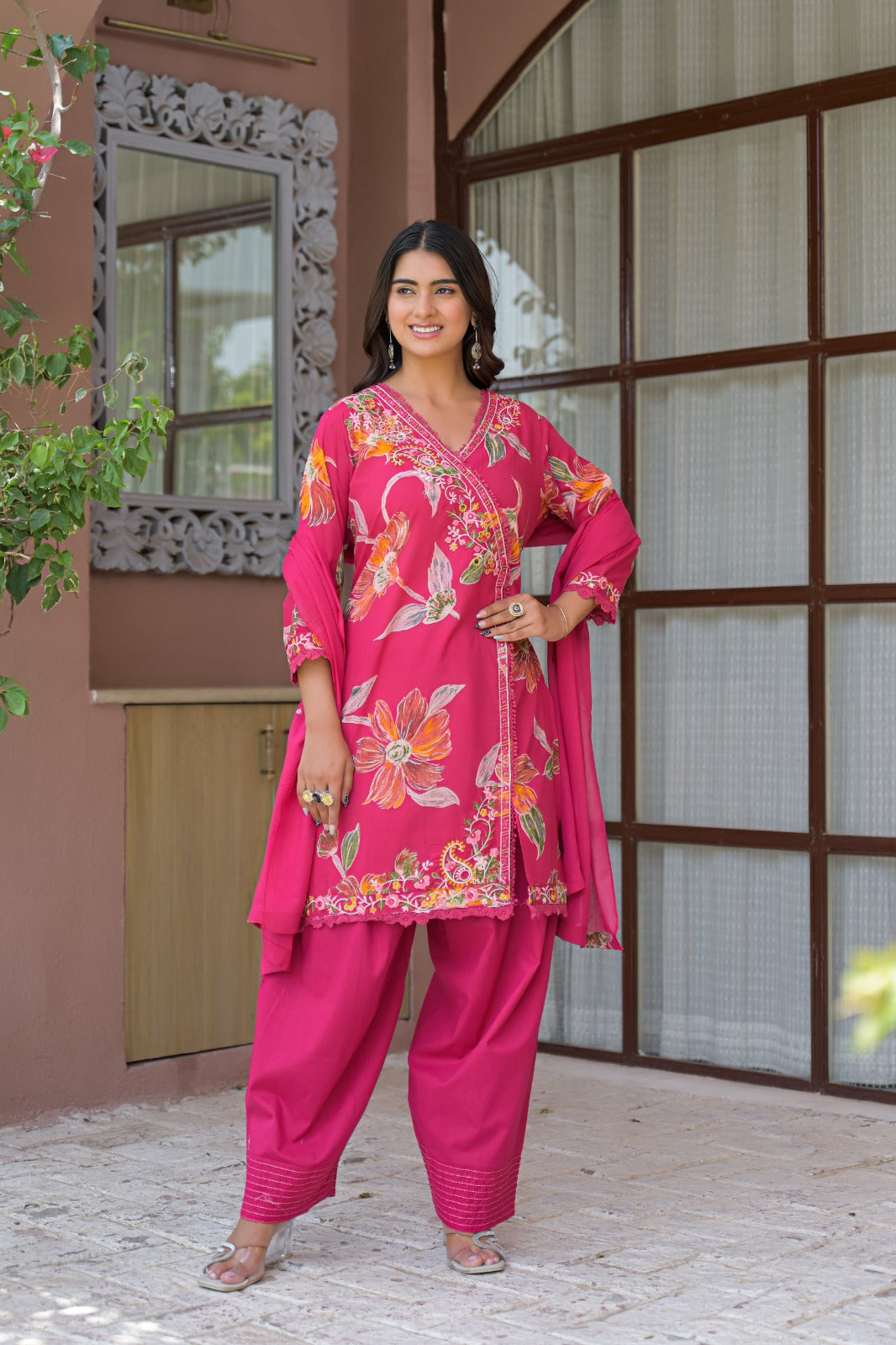 A woman modeling a bright pink Indian ethnic salwar kameez suit with floral embroidery and a matching dupatta, standing outdoors in front of a building with large window panes.