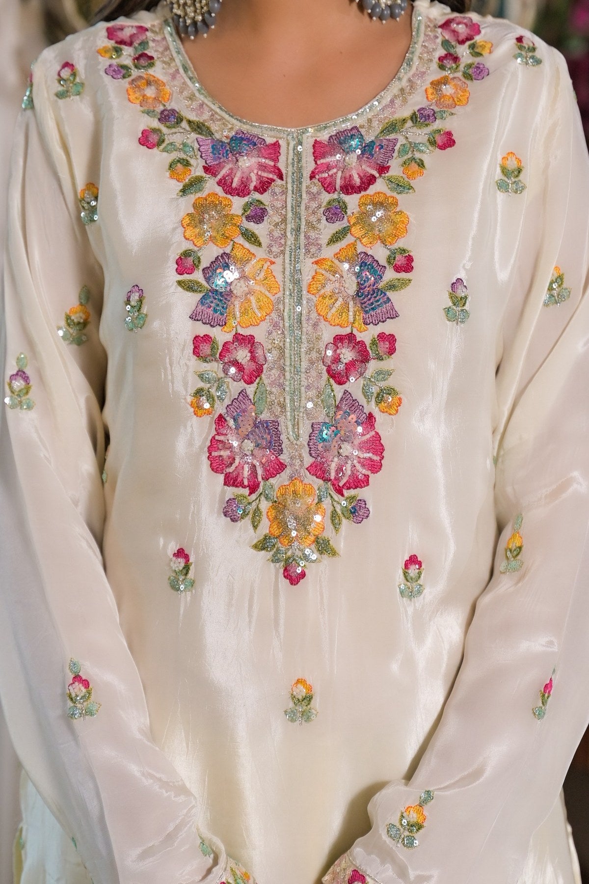 A close-up of a cream-colored embroidered traditional garment, likely a kurta, featuring a colorful floral pattern with pink, yellow, and purple flowers and green leaves on the chest and scattered on the sleeves.