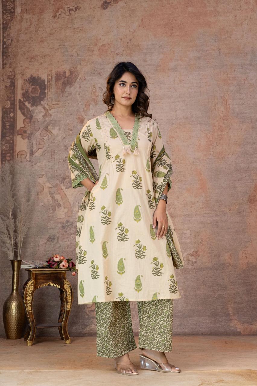 A woman models a traditional Indian outfit consisting of a cream-colored kurta with green leaf prints and green patterned sleeves, paired with matching green patterned pants, set against a rustic textured wall backdrop.