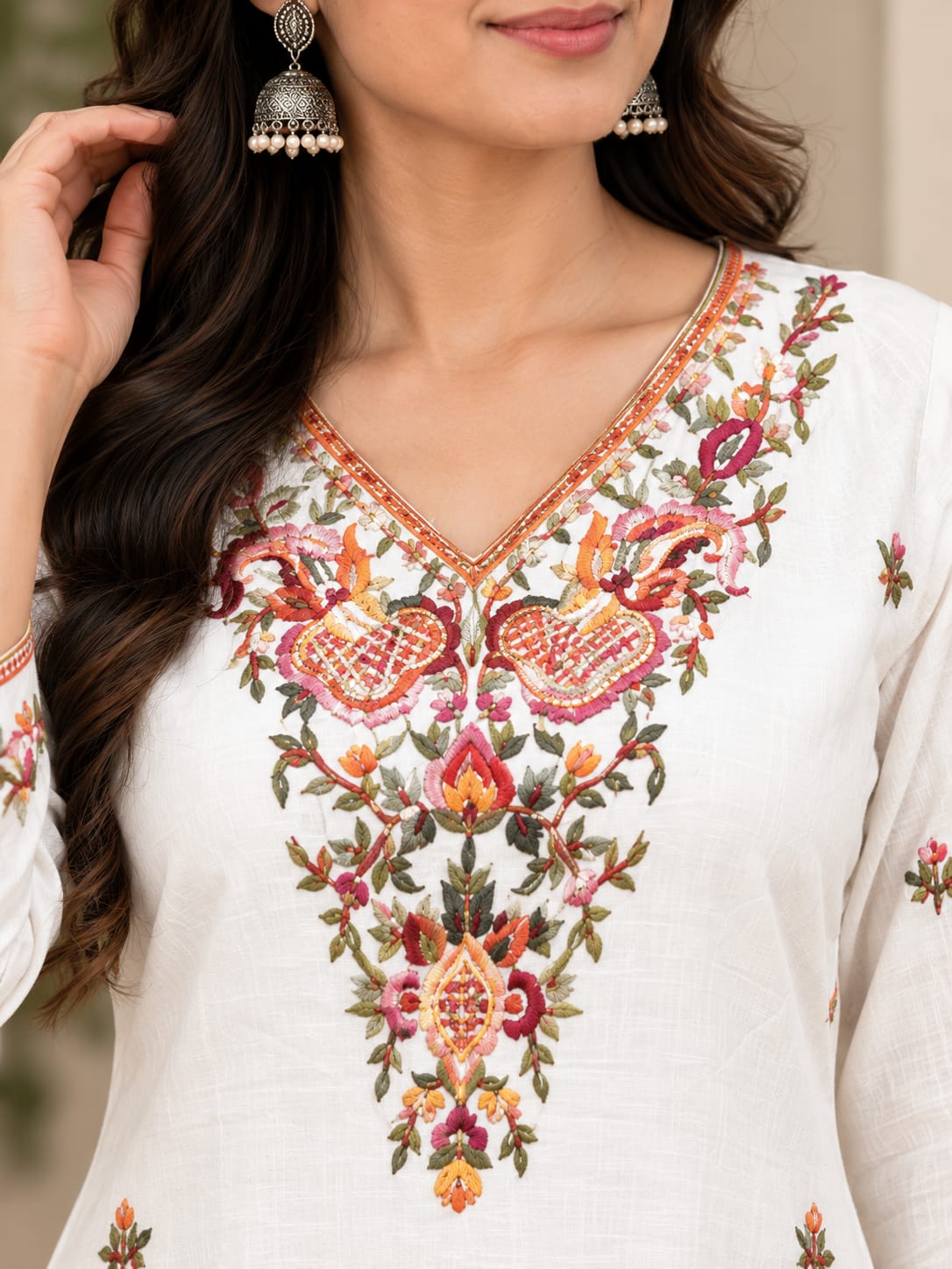 A close-up of a woman wearing a white kurta with intricate multicolor floral embroidery around the V-neckline. She is also wearing silver jhumka earrings, and the background is a softly blurred neutral indoor setting.