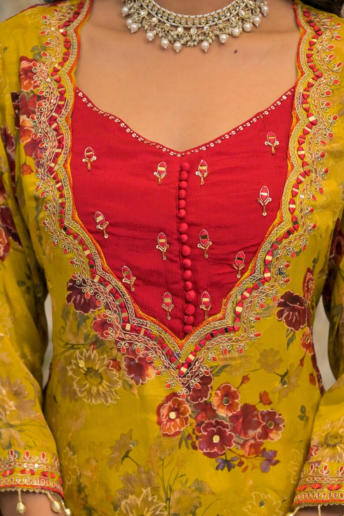 Close-up of a traditional Indian outfit featuring a vibrant yellow base with a red V-neck panel, detailed floral embroidery, and a silver-toned pearl necklace.