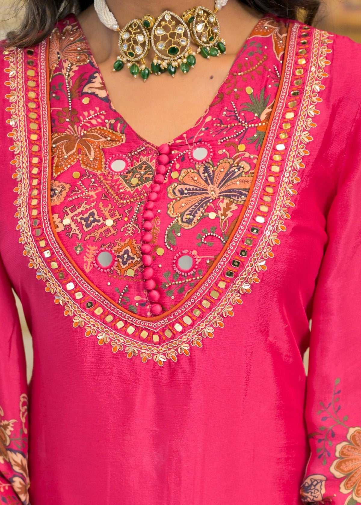 A close-up of a bright pink traditional embroidered garment with a V-neckline, featuring intricate floral and geometric embroidery in gold, green, and brown, adorned with mirror work and bead details.