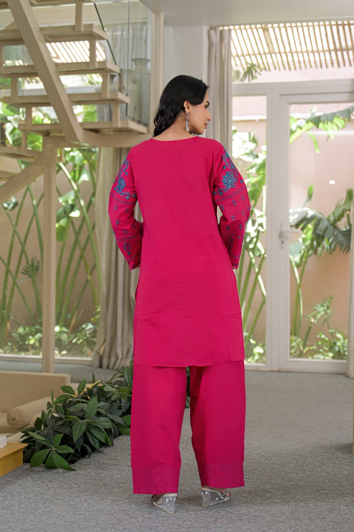 A woman stands with her back to the camera wearing a bright pink kurta and trouser set with teal embroidery on the sleeves, set in a modern indoor space with plants and a staircase.