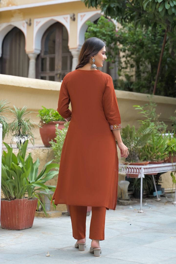 A woman wearing a rust-orange kurta and matching trouser set, viewed from behind in an outdoor courtyard setting with potted plants and a building in the background.
