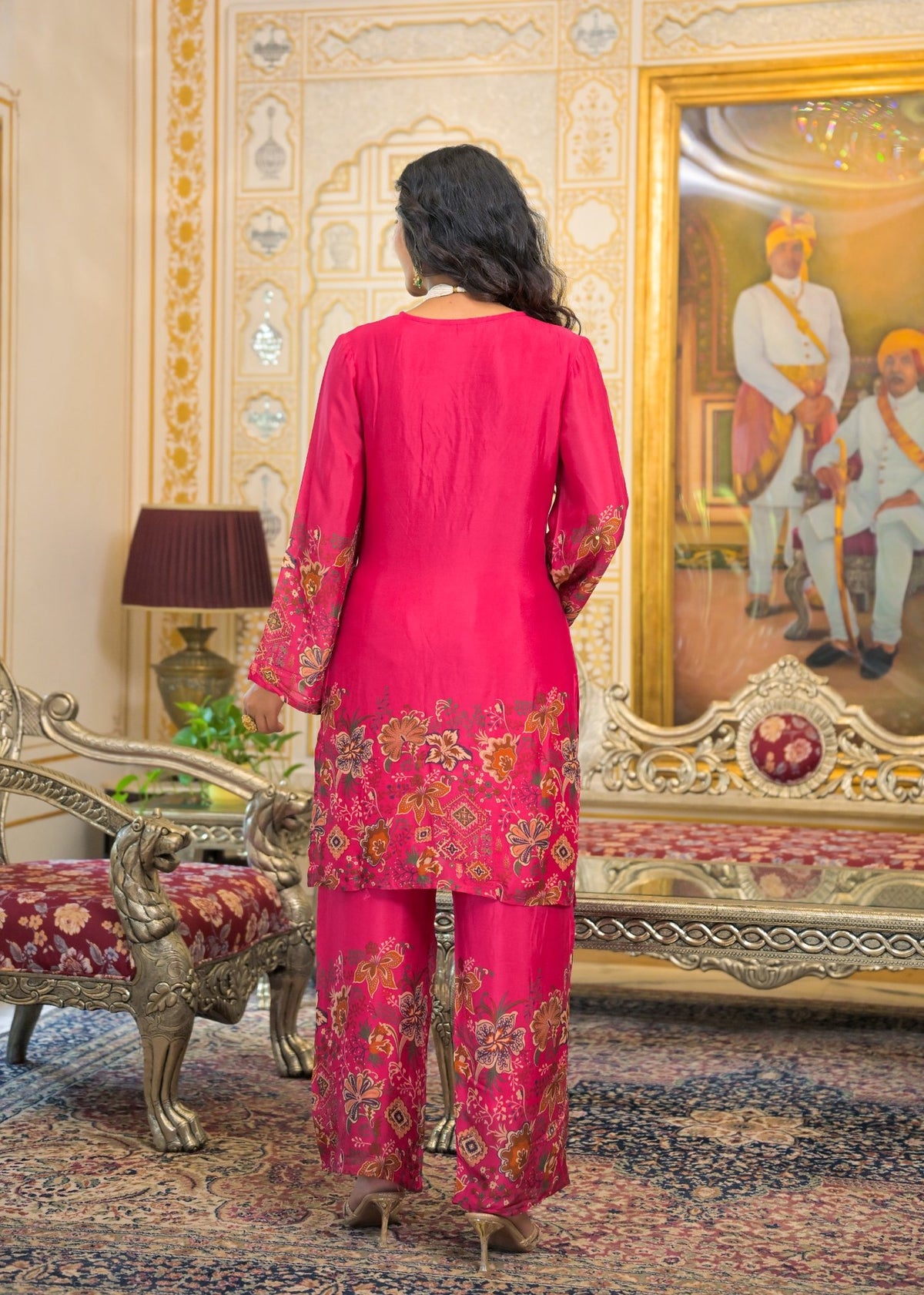 A woman viewed from behind wearing a bright pink traditional outfit with floral embroidery, standing in an ornate room with a large framed painting and decorative furniture.