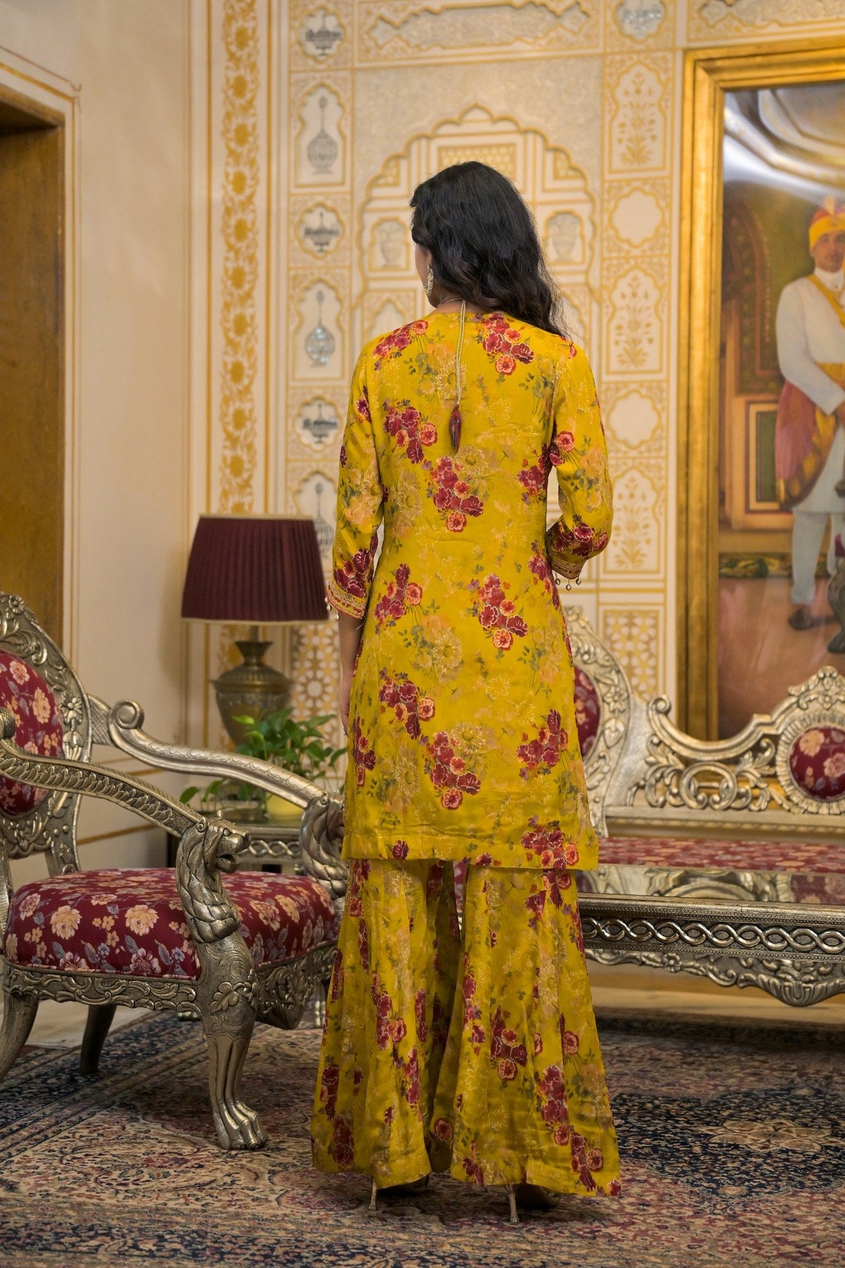 A woman stands with her back to the camera wearing a yellow three-piece salwar kameez suit with a red floral print, set in an ornate traditional room with gold-patterned walls and carved furniture.