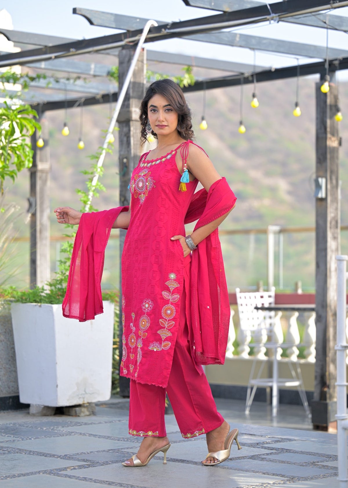 A woman models a bright pink traditional Indian salwar kameez suit with gold embroidery and a matching dupatta in an outdoor patio setting.