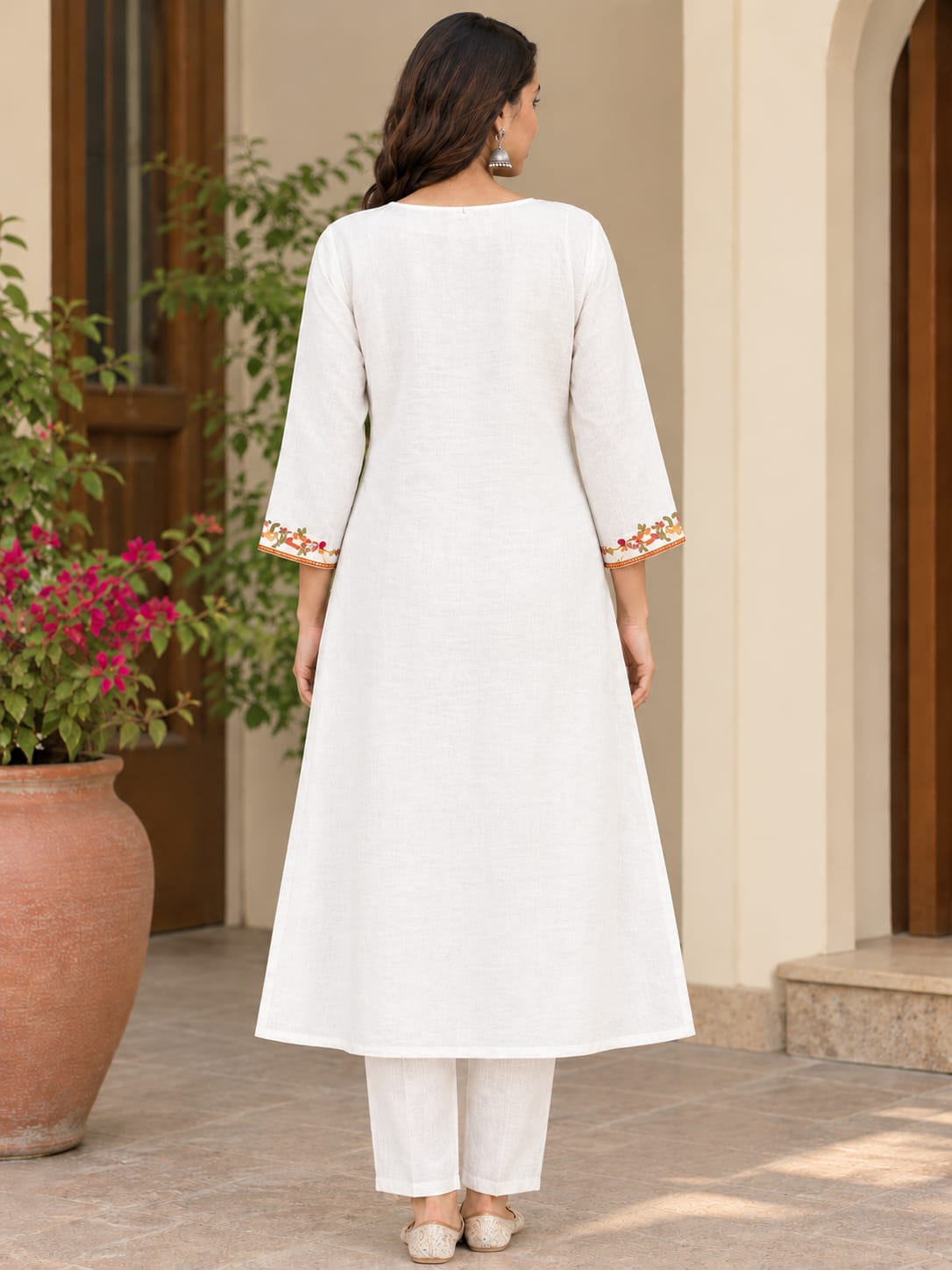 A woman viewed from behind wearing a white A-line kurta with three-quarter sleeves and colorful floral embroidery on the cuffs, paired with white trousers, set against a warm beige architectural background with a potted plant.