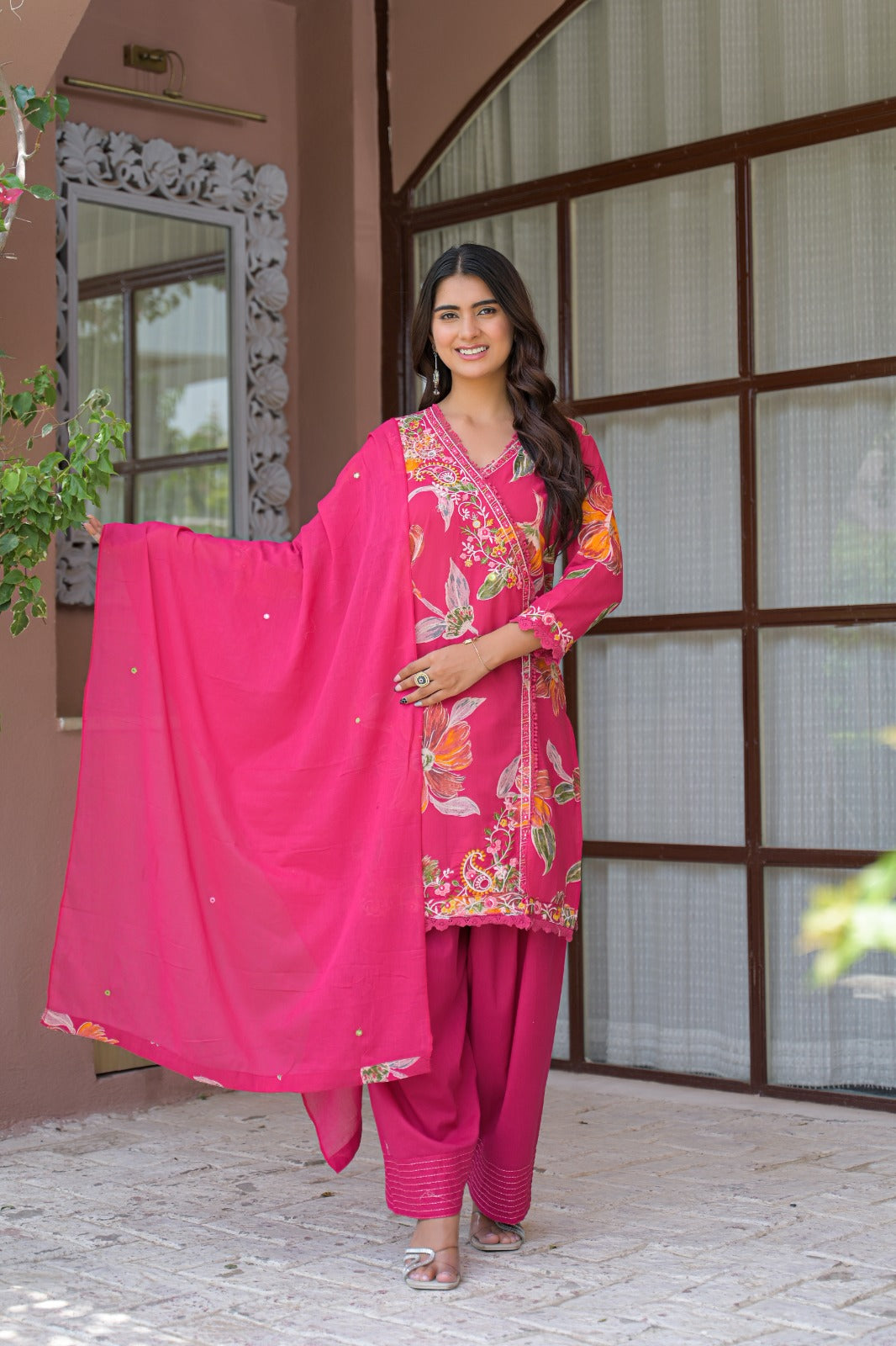A woman modeling a bright pink Indian ethnic salwar kameez suit with floral embroidery and a matching dupatta, standing outdoors in front of a building with large window panes.