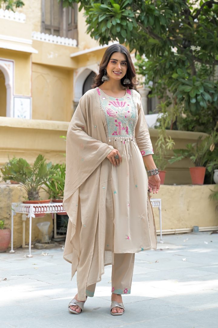 A woman models a beige Indian ethnic outfit consisting of a kurta with pink and green floral embroidery, matching trousers, and a draped dupatta, set against a warm yellow building background.