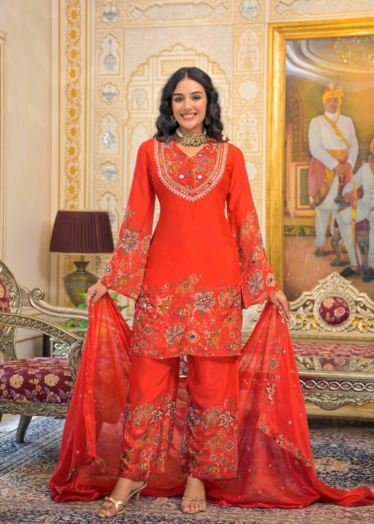 A woman models a traditional red Indian salwar kameez suit with intricate gold and silver embroidery, standing in an ornate, vintage-style room with a large painting and carved furniture.
