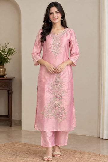 A woman modeling a light pink traditional Indian salwar kameez set with intricate silver and gold embroidery, standing in a neutral indoor setting.