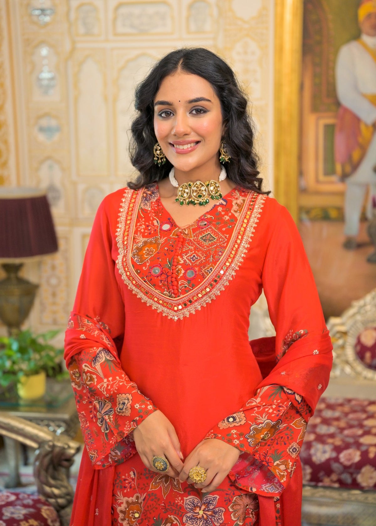 A woman wearing a bright red traditional Indian salwar kameez outfit with intricate gold and multicolor embroidery, standing in an ornate indoor setting.
