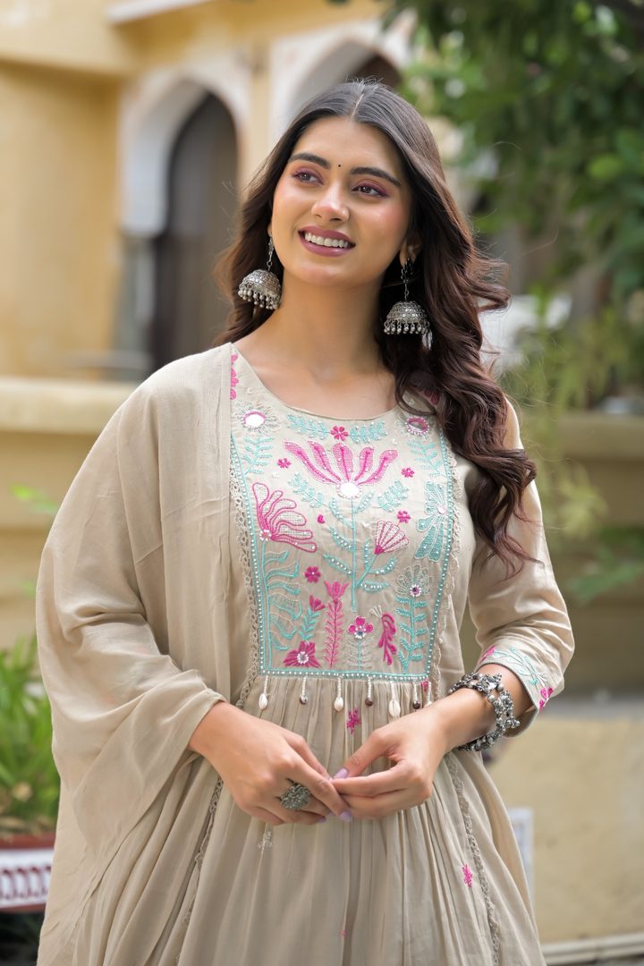 A woman wearing a beige traditional outfit with a light blue and pink embroidered bodice, standing outdoors in front of a beige building with green foliage.