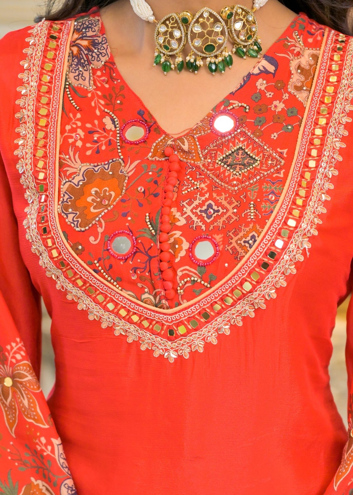 A close-up of a bright orange-red traditional embroidered top with a large V-neckline, decorated with intricate gold, green, and multicolor floral and geometric patterns, paired with a green beaded necklace.