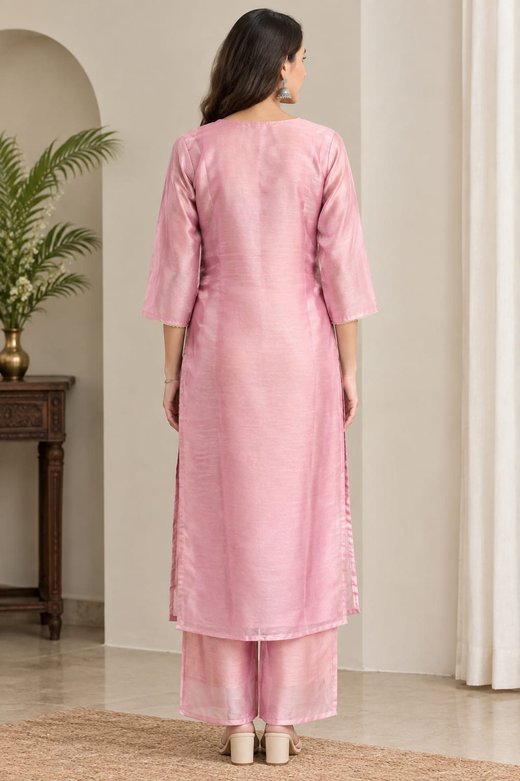 A woman viewed from behind wearing a light pink three-quarter sleeve kurta and matching trousers, standing in an indoor room with neutral walls, a potted plant, and a curtain.