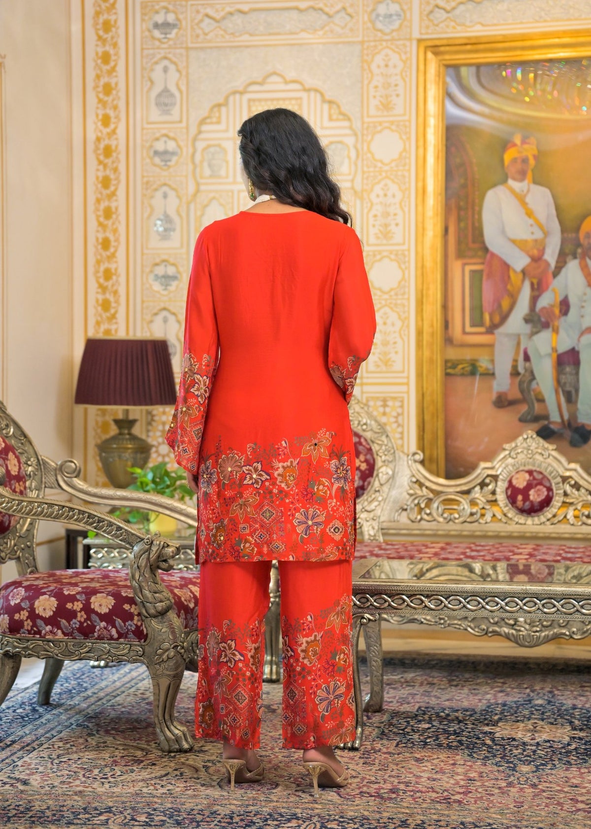 A woman viewed from behind wearing a bright red traditional outfit with ornate floral embroidery concentrated along the hem and lower pant legs, standing in an ornate interior with carved furniture and a large framed painting.