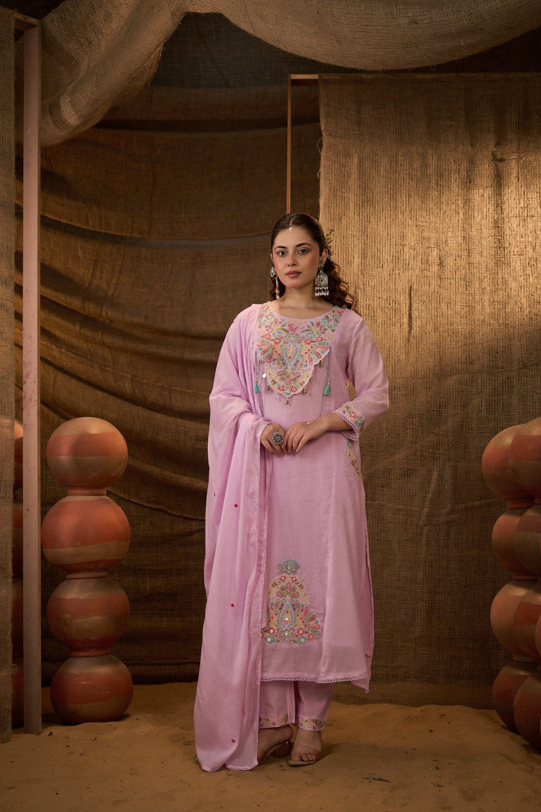 A woman stands in a rustic indoor setting wearing a light pink traditional outfit with colorful embroidered details on the chest and lower kurta, paired with a matching dupatta. The background features a burlap curtain and stacked terracotta pots.