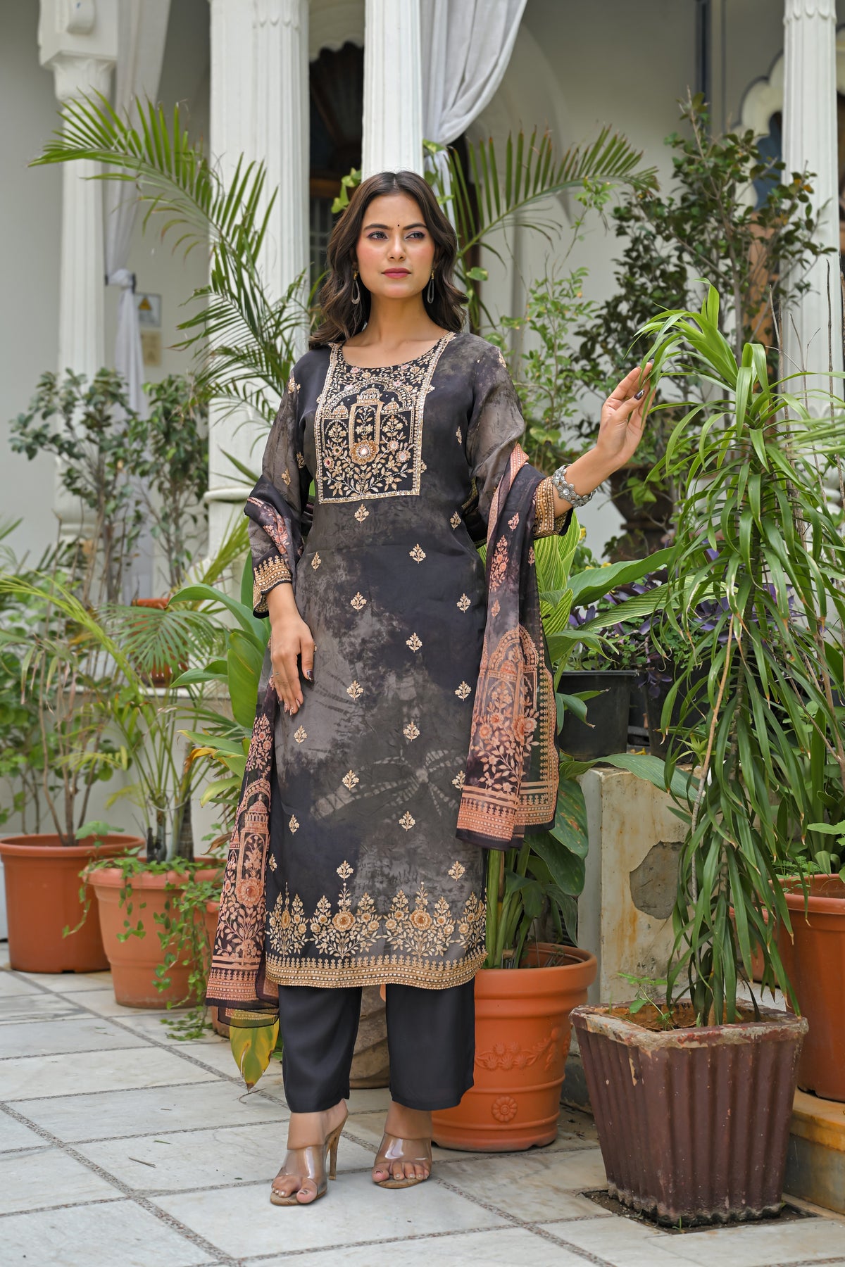 A woman models a dark gray and black printed salwar kameez suit with gold embroidery, black trousers, and a patterned dupatta in an outdoor patio setting with potted plants.