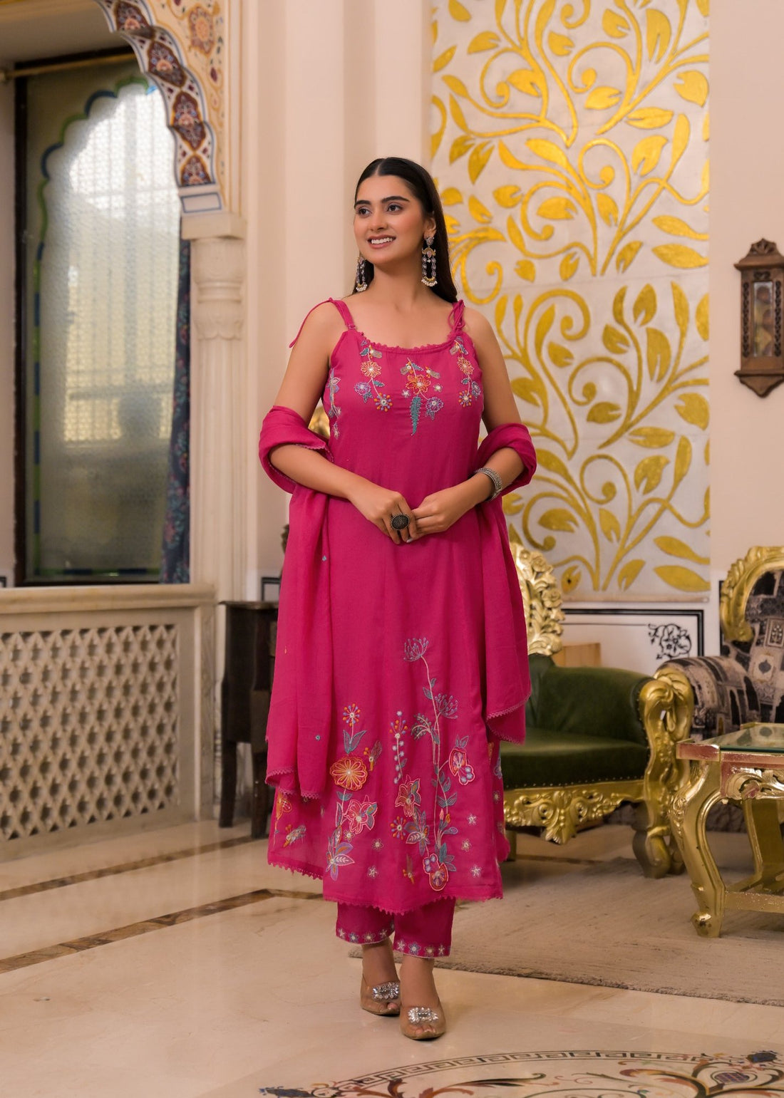 A woman modeling a bright pink embroidered kurta set with a dupatta in an ornate indoor setting with gold-patterned walls and green-gold furniture.