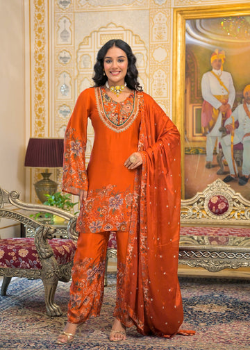 A woman models an orange traditional Indian salwar kameez suit with floral embroidery and a matching dupatta in an ornate, vintage-style room with gold-framed paintings and patterned furniture.