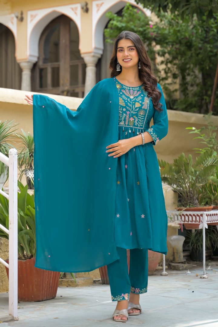 A woman models a teal blue ethnic salwar kameez suit with intricate embroidery, featuring a matching dupatta, in an outdoor courtyard setting.
