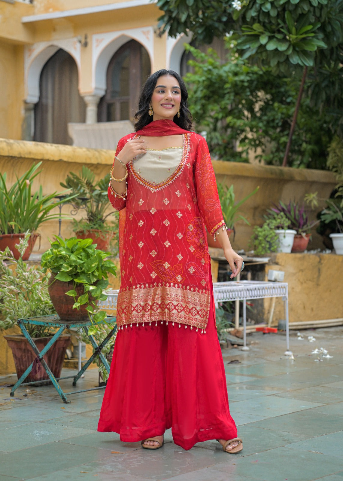 A woman stands outdoors wearing a bright red traditional outfit with a matching dupatta, featuring gold embroidery and a beige inner top. The setting includes potted plants, a stone patio, and a yellow building in the background.
